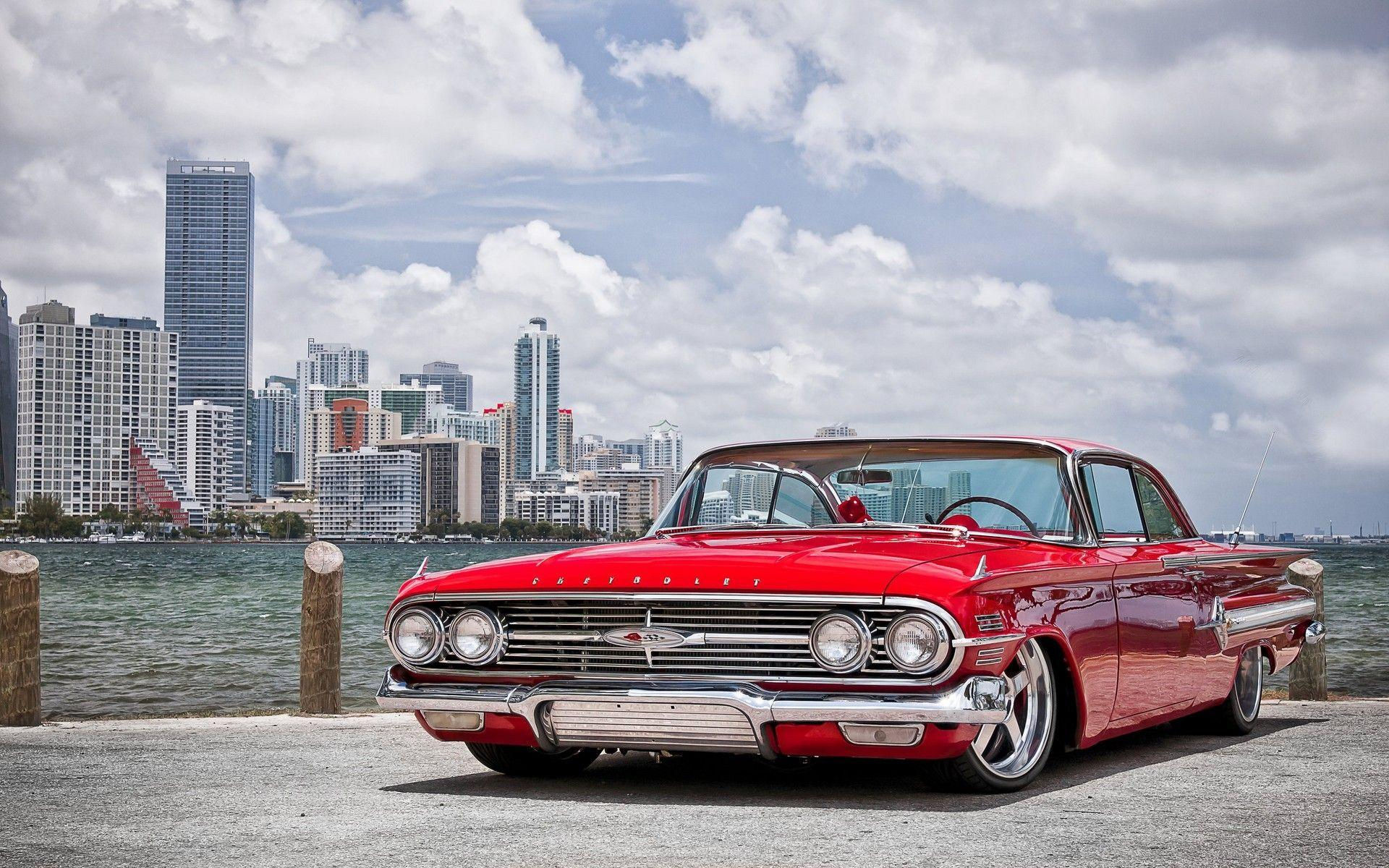 Wallpaper #_3MOho4BFI5NbQksfx5p0 A Red Chevrolet Impala Parked by the Water with a Cityscape in the Background