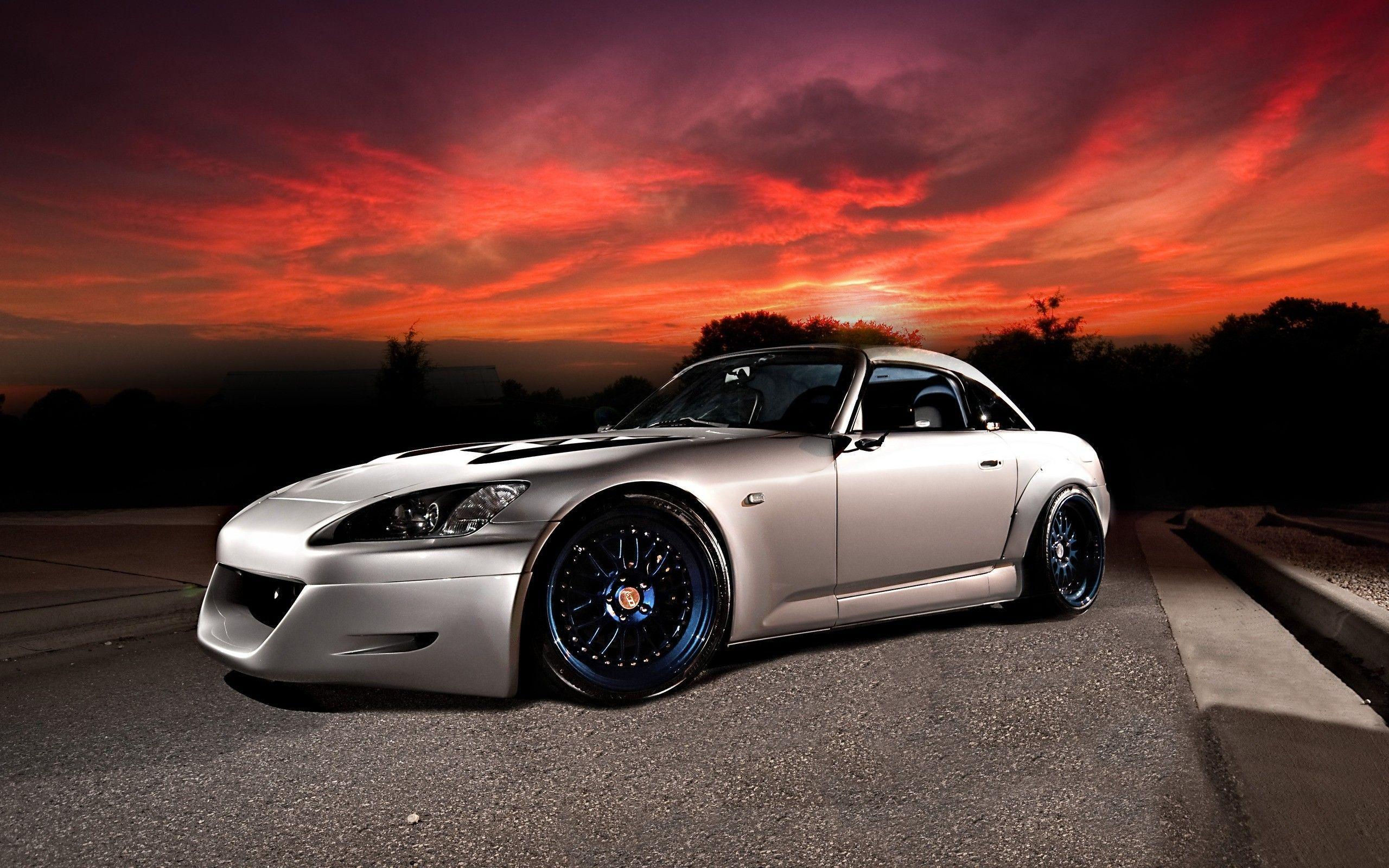 Wallpaper #r0U0l44B7YBJg1BVZKDq10 A Silver Sports Car Parked on a Road with a Red and Orange Sky in the Background