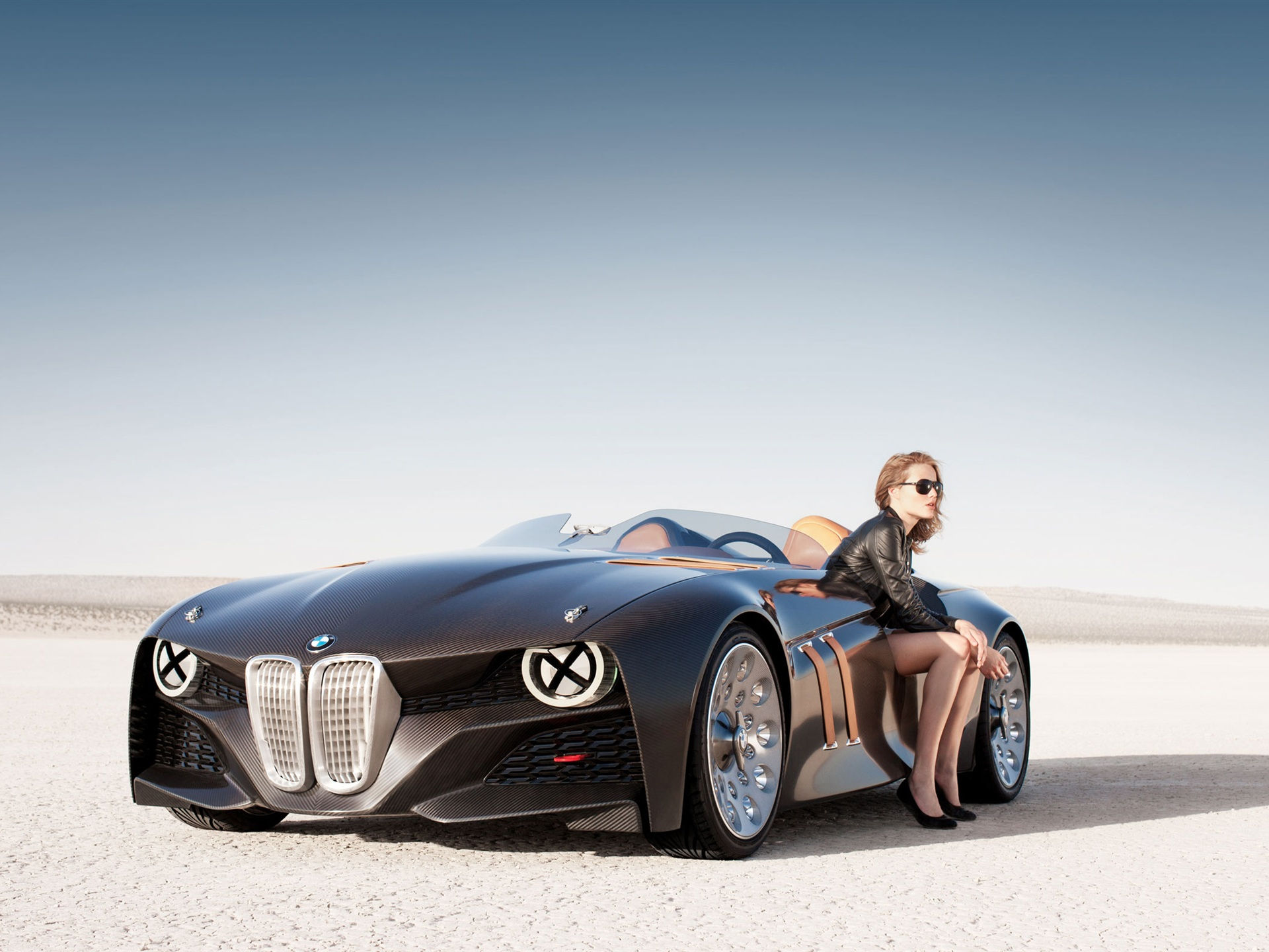 Wallpaper #w0Xoko4B7YBJg1BVD55I8 A Woman in a Black Leather Jacket Poses on the Door of a Brown BMW Concept Car in the Desert